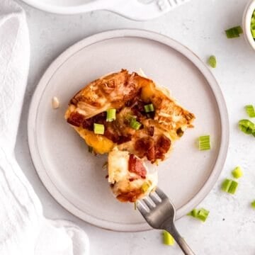closeup overhead square hero shot of a slice of savory bacon swiss croissant breakfast casserole on a white earthenware plate with a silver fork taking a bite out.
