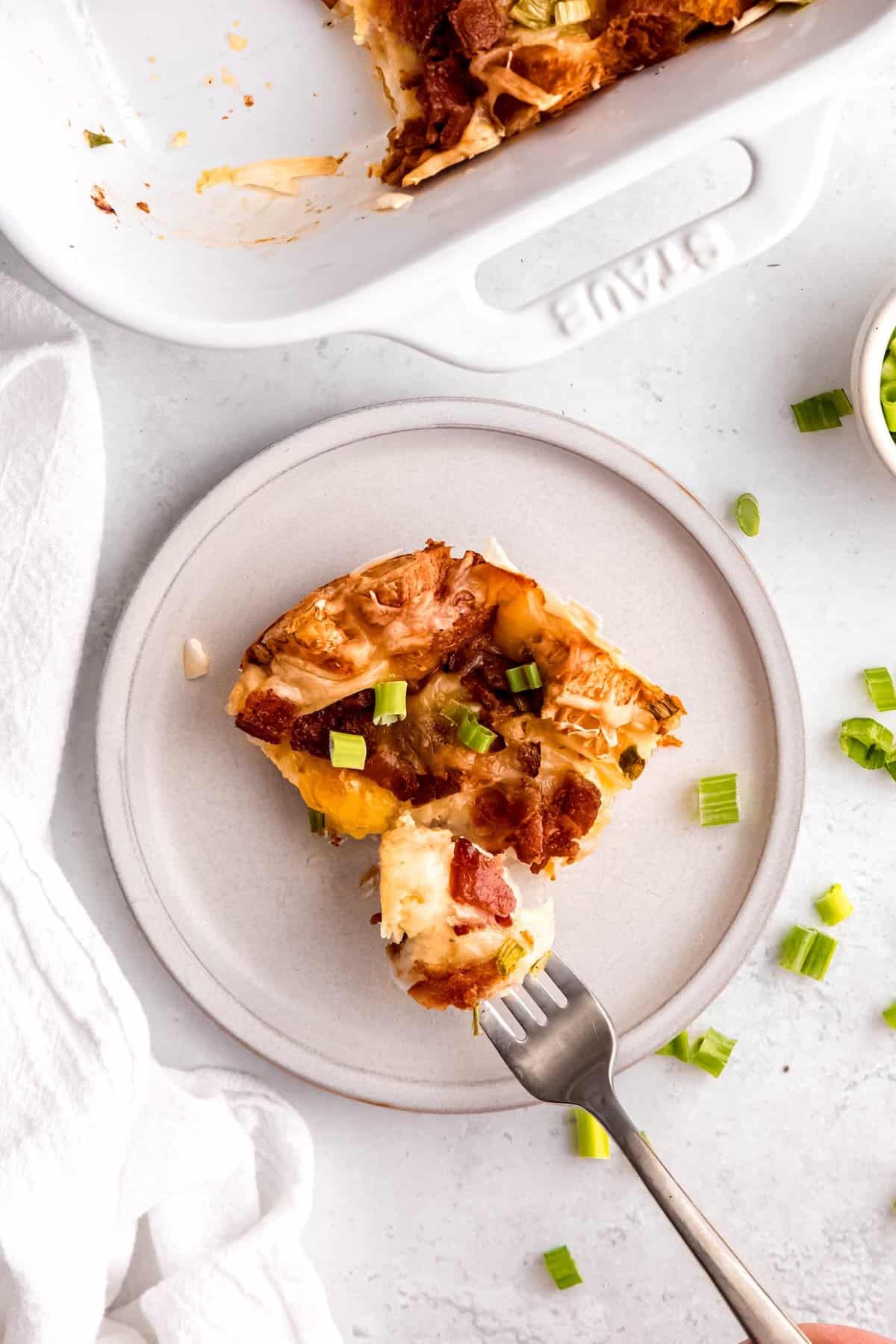 silver fork taking a bite of a slice of savory bacon swiss breakfast strata made with croissants, showing the casserole layers of meat, cheese, fluffy croissants, and fresh scallions.