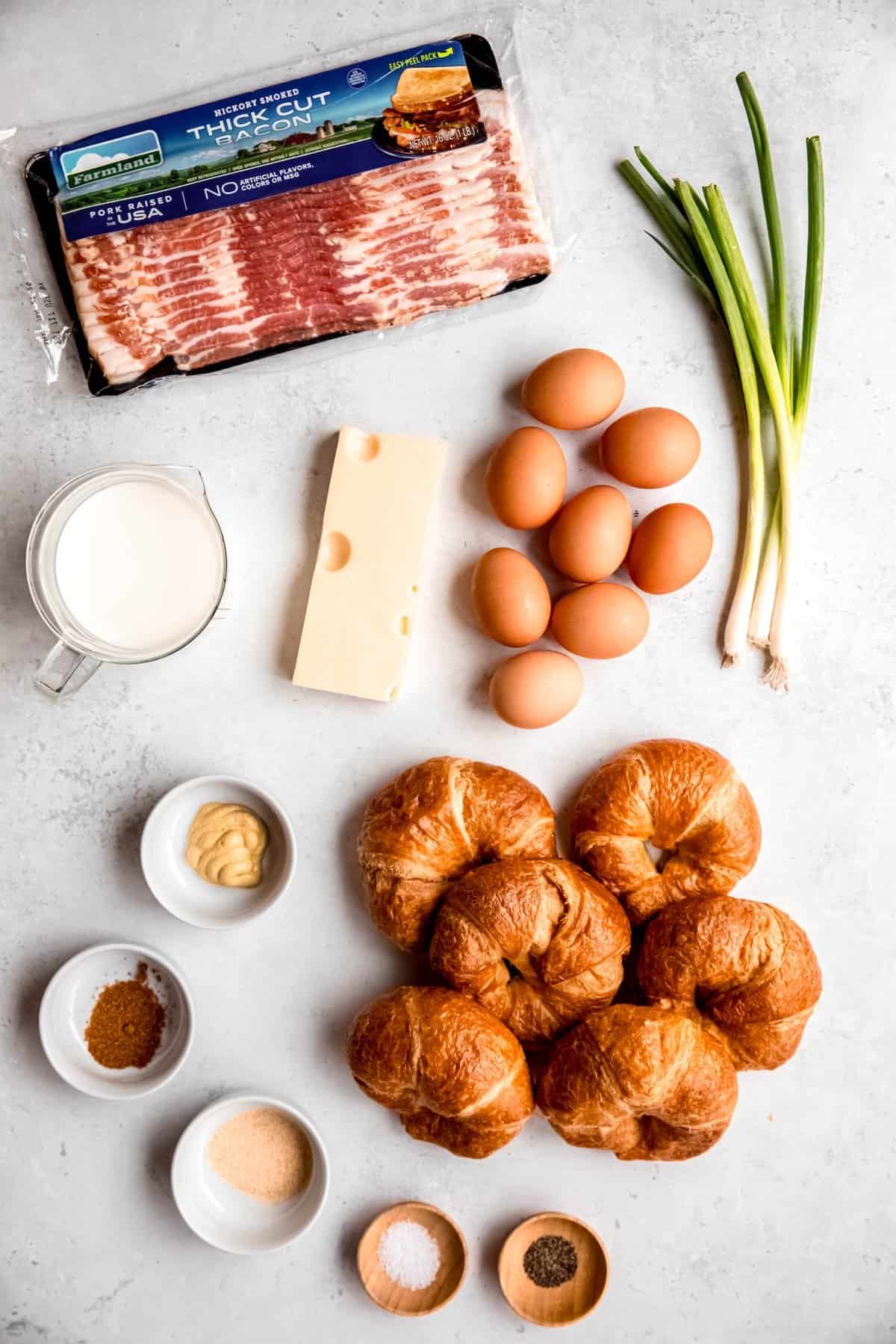 ingredients needed to make a savory croissant breakfast casserole recipe with bacon and swiss measured out on a white table.