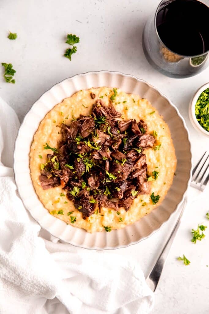 Italian braised beef short rib polenta dinner served in a white pasta bowl with a glass of red wine.