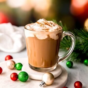 clear mug filled with spiked christmas coffee topped with amaretto whipped cream on a white table with red, white and green christmas ornaments and a sprig of pine in front of blurred out christmas lights.