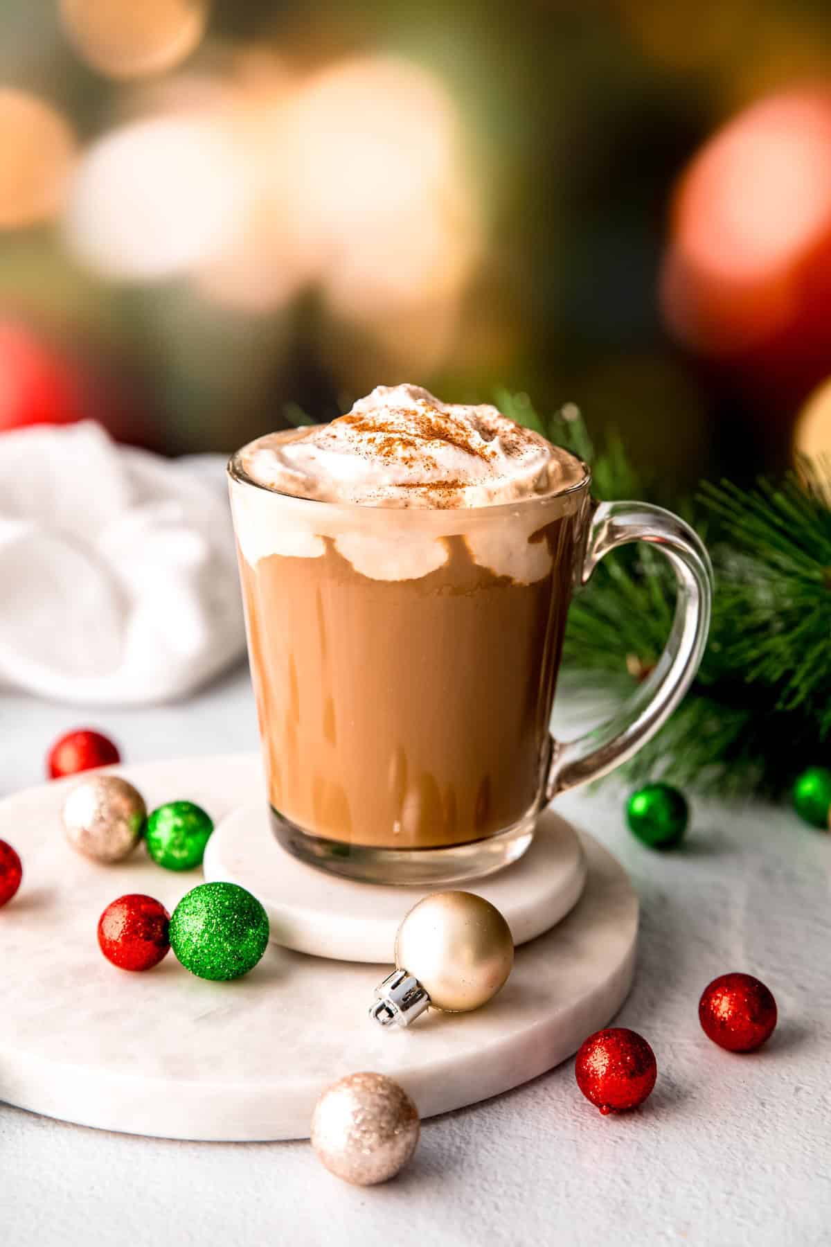 clear mug filled with spiked christmas coffee topped with amaretto whipped cream on a white table with red, white and green christmas ornaments and a sprig of pine in front of blurred out christmas lights.
