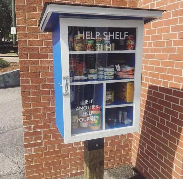 small community food box set up in front of a library with the words "help shelf" "help another, help yourself" on the front.