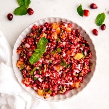 square hero image of a bowl of orange cranberry jalapeno relish/salsa/dip on a white table with fresh mint leaves and fresh cranberries.