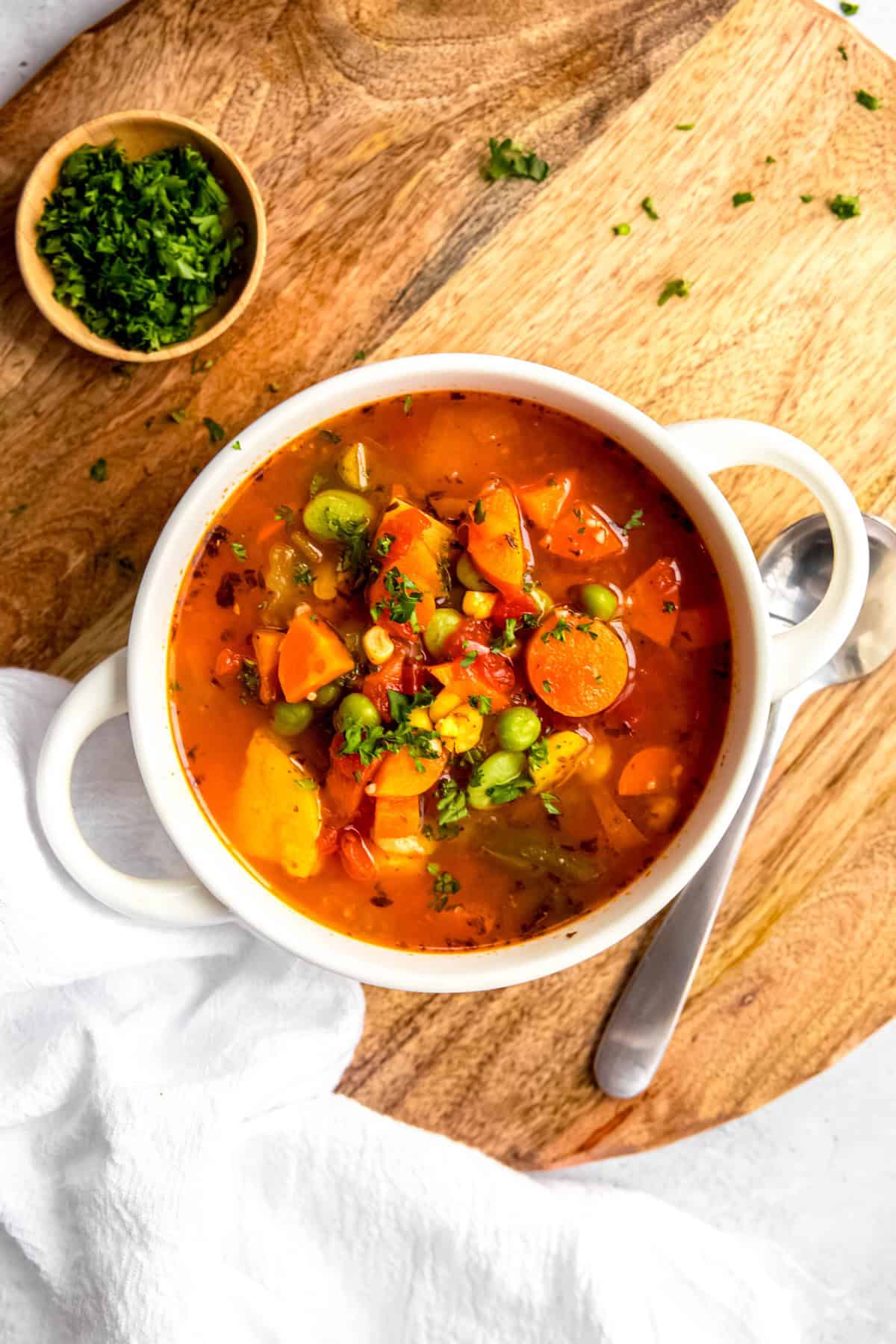 flat lay shot of a serving of easy instant pot vegetable soup in a white bowl with handles on a wooden round trivet with a silver spoon, a pinch bowl of fresh parsley, and a white linen napkin.