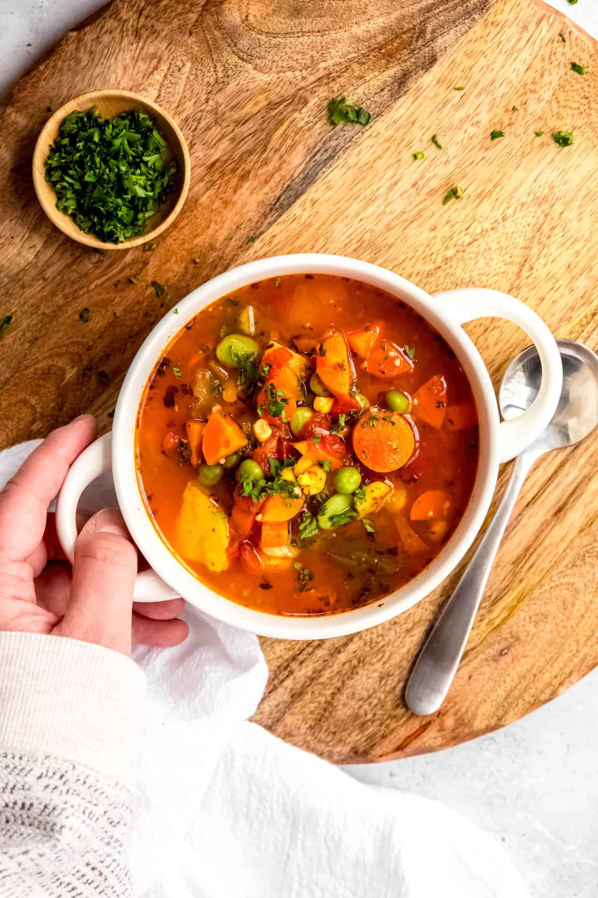 hand in a white sweater up to the knuckles setting down a bowl of instant pot vegetable soup on a wooden trivet with a silver spoon and a bowl of fresh parsley.