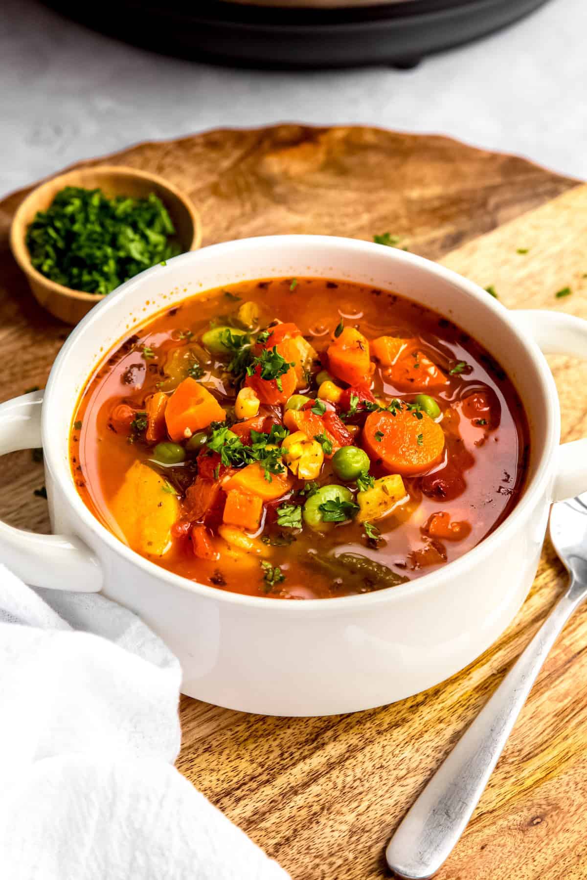 45 degree angle shot of a white bowl with handles filled with instant pot vegetable soup made with frozen vegetables on a wooden trivet with a wooden pinch bowl of chopped parsley and a silver spoon.