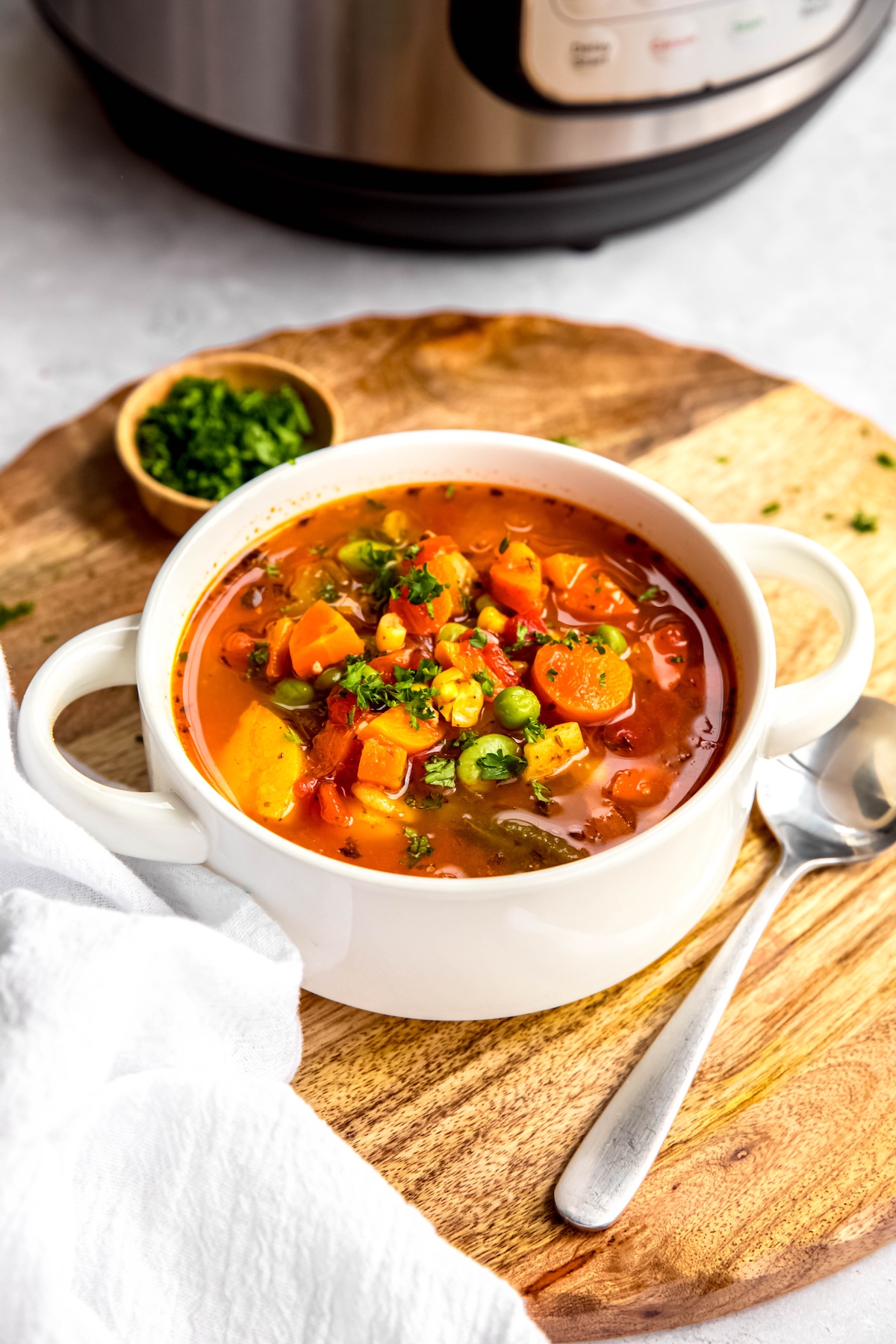 45 degree angle shot of a serving of vegetable soup made with frozen veggies on a wooden trivet with a silver spoon and a pinchbowl of fresh parsley and an instant pot blurred out in the background.
