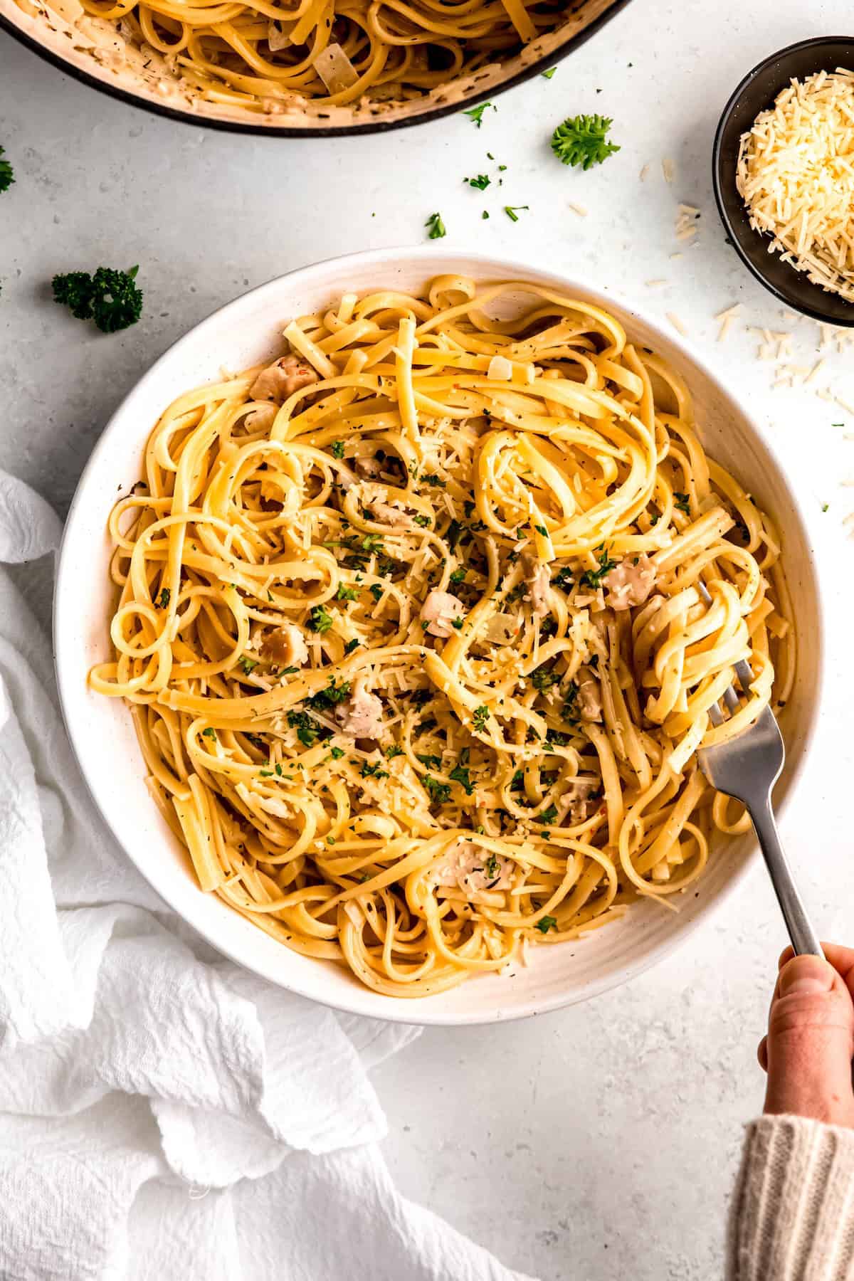 overhead shot of a white pasta bowl filled with canned clam linguine in white wine sauce garnished with fresh parsley and grated parmesan on a white table; a hand in a white sweater is twirling up a bite on a silver fork.
