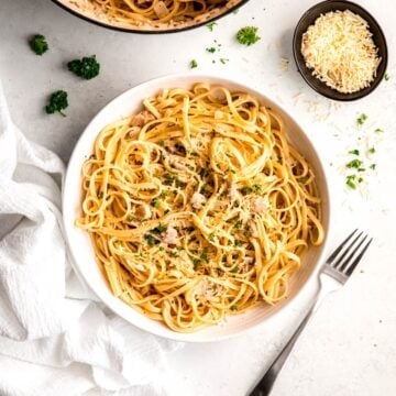 square hero of a plate of canned clam linguine with creamy white wine sauce on a white table with a silver fork, fresh parsley, and a black pinch bowl of grated parmesan.