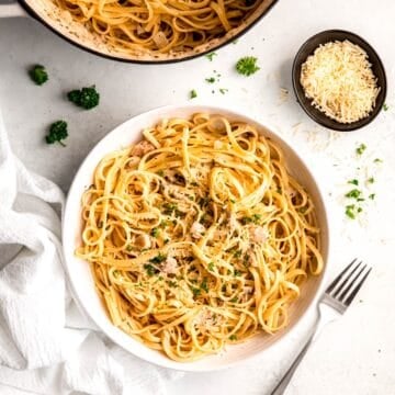 overhead shot of a white table with a pot of easy canned clam linguine with a white pasta bowl filled with a portion of it topped with fresh parsley and cheese.