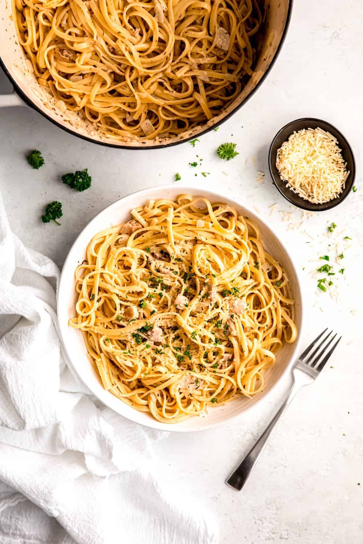 overhead shot of a white table with a pot of easy canned clam linguine with a white pasta bowl filled with a portion of it topped with fresh parsley and cheese.