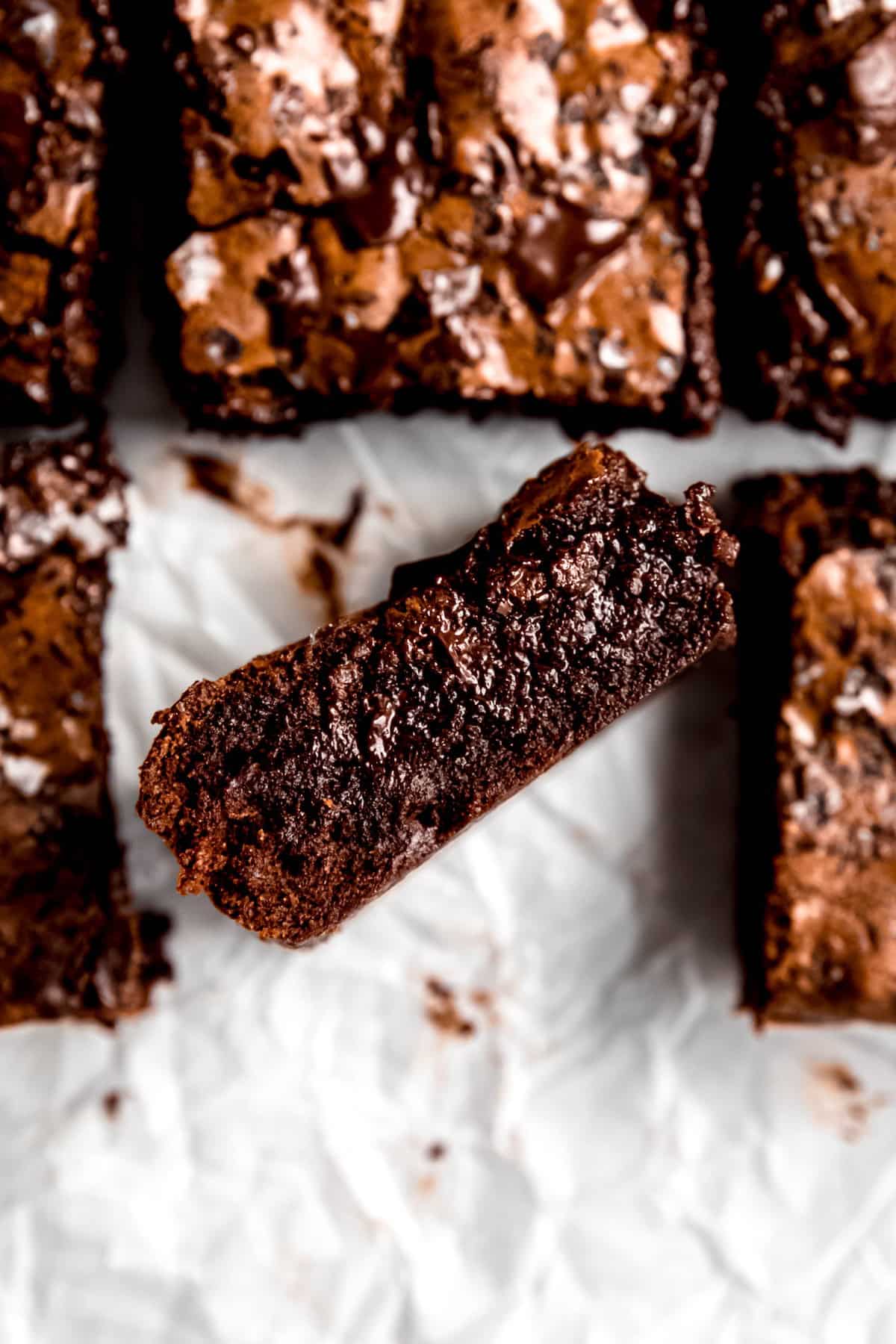 overhead shot of a tray of gooey dark chocolate brownies with one turned on its side to show the super fudgy interior of the brownies while the shiny, crinkly tops of the remaining brownies are slightly blurred out below.