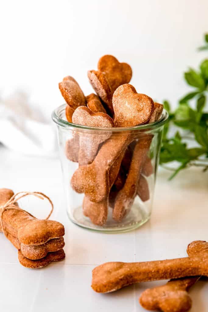clear jar of homemade banana oat dog treats with peanut butter powder on a white table with another stack of three bones tied together with twine and a green houseplant.