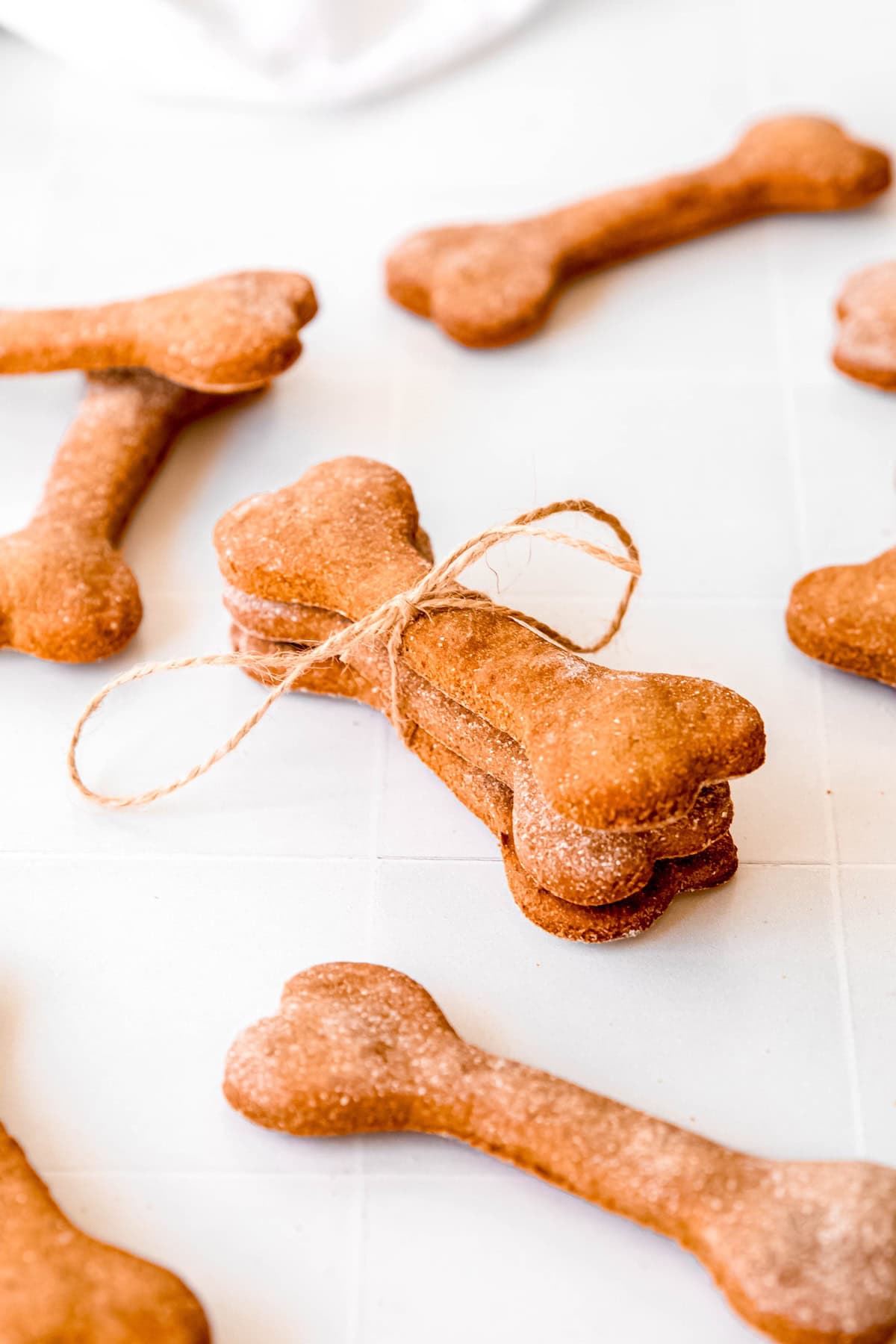 stack of 3 homemade oat banana and peanut butter dog treats that are shaped like bones tied with natural twine on a white table with more dog biscuits scattered around.