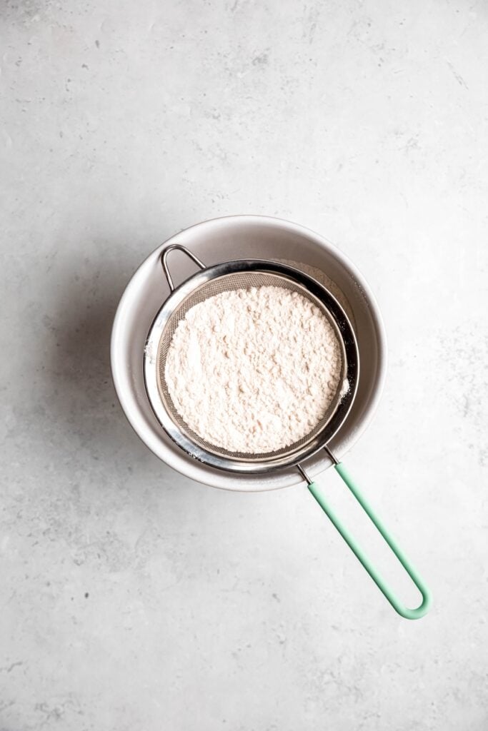 flour and salt being sifted together into a bowl using a mesh strainer.