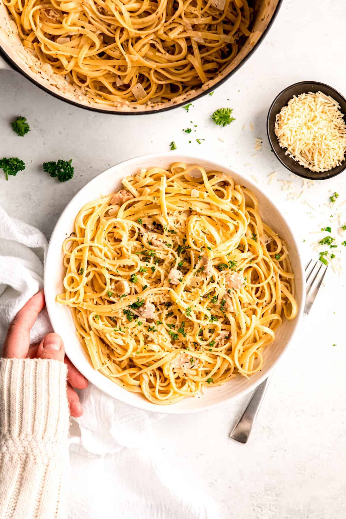 hand in a white sweater up to the knuckles setting a white pasta bowl of linguine in clam sauce with canned clams down on a white table with a silver fork with the rest of the dutch oven filled with the batch and a bowl of grated parmesan.