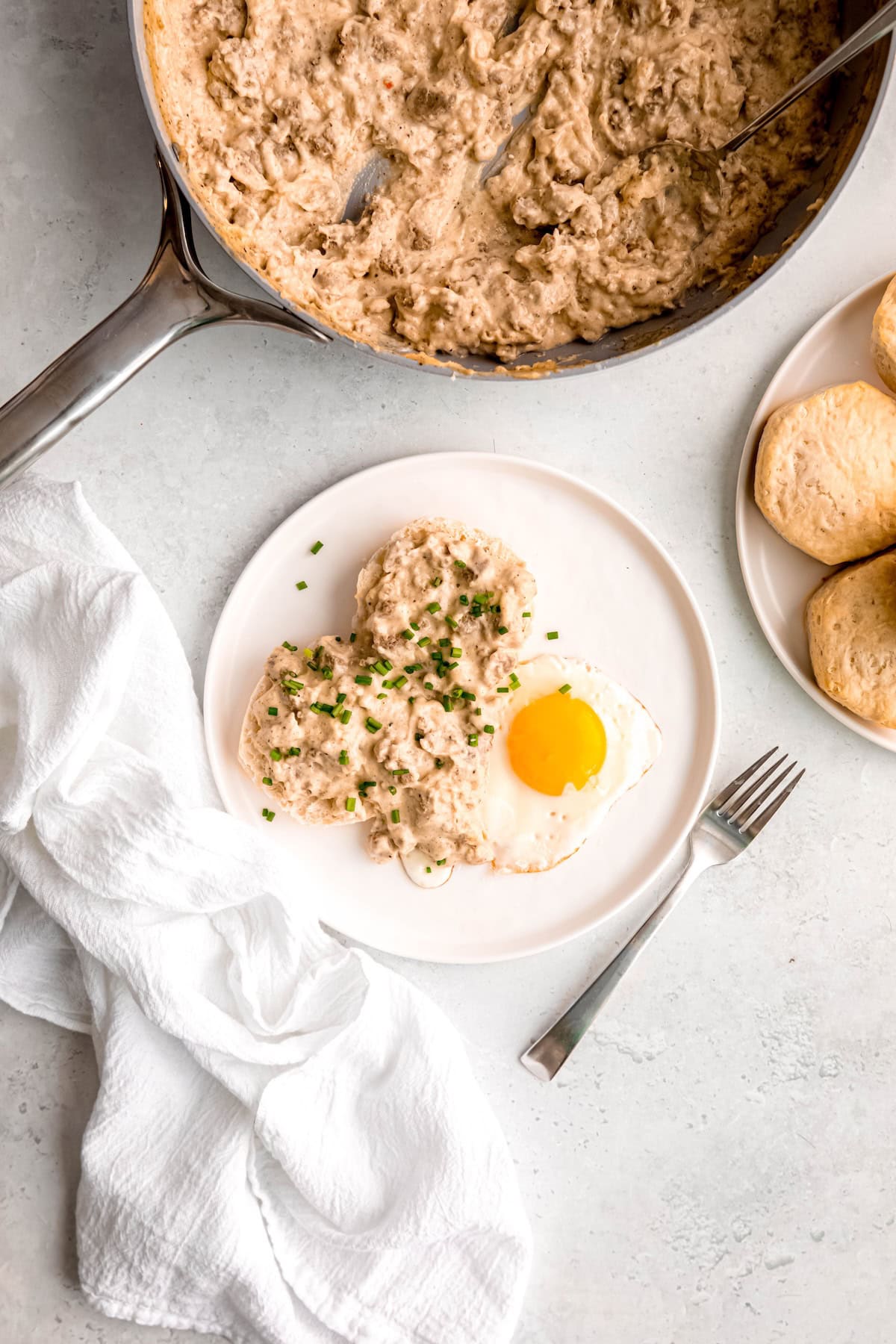 overhead shot of a white table with a white plate with a split biscuit topped with white sausage gravy garnished with fresh chives and a sunny side egg with a plate with more biscuits and the skillet with the rest of the country gravy framing the shot.