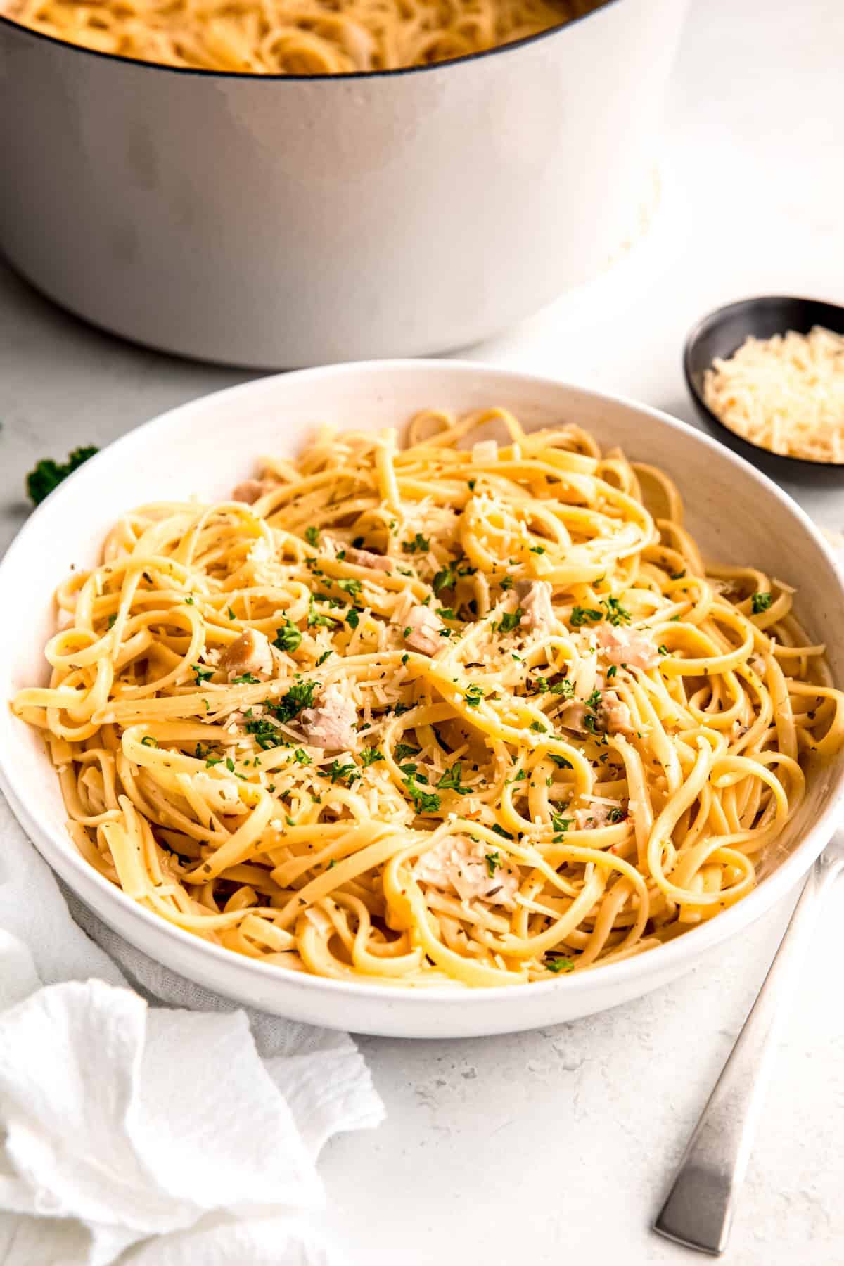 white pasta bowl of pasta in white clam sauce topped with fresh parsley and parmesan cheese on a white table with a silver fork with a black pinch bowl of parmesan cheese and a white dutch oven with the rest of the linguine & clams in the background.