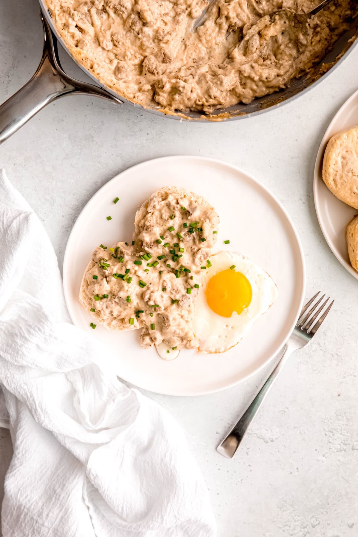 overhead shot of sausage gravy and biscuits plated up with a sunny side up egg and fresh chives on a white plate with a silver fork.