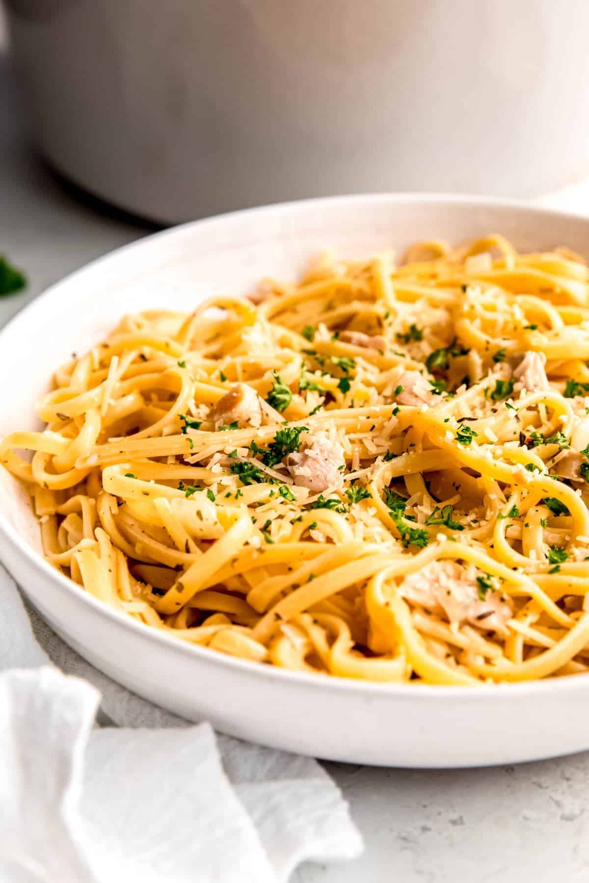 45 degree angle closeup of a white pasta bowl filled with a portion of white clam sauce canned clam pasta garnished with fresh parsley and grated parmesan cheese in front of a white dutch oven on a white table with a white linen.