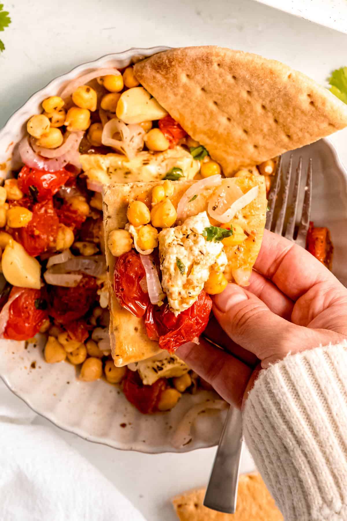 hand in a white sweater up to the knuckles scooping up a bite of baked feta, tomatoes and chickpeas on a wedge of flatbread.