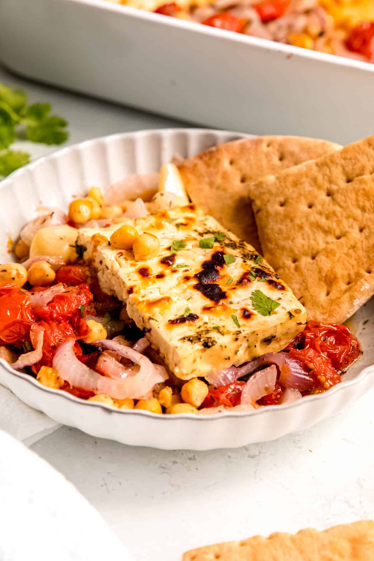 45 degree angle shot of a scalloped white pasta bowl filled with a portion of baked feta with tomatoes and chickpeas and two wedges of pita bread.