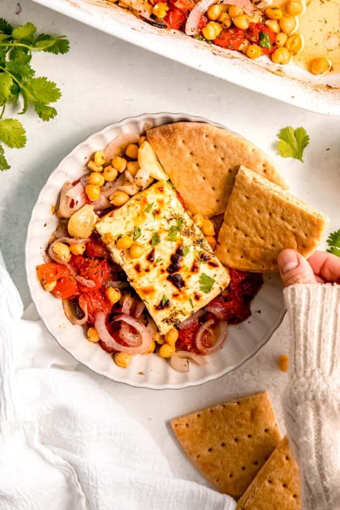 hand clad in a cozy white sweater picking up a quarter of pita bread to dip into the baked feta with tomatoes and chickpeas.
