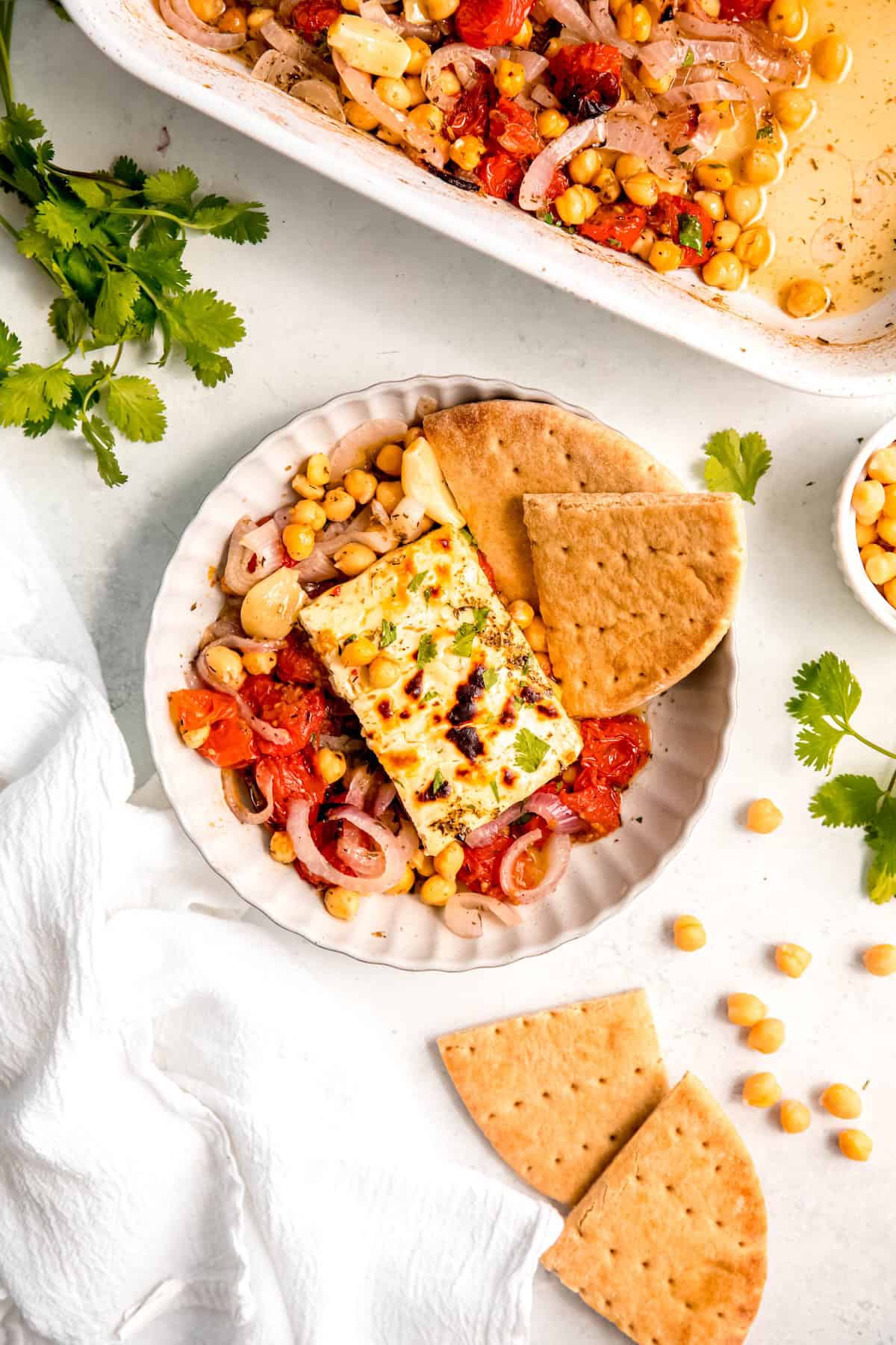 overhead shot of a white scalloped pasta bowl filled with a portion of baked feta with tomatoes and chickpeas on a white table with the remaining recipe in the baking dish framing the top of the shot, a couple of bunches of fresh cilantro to either side, and a few wedges of pita bread.