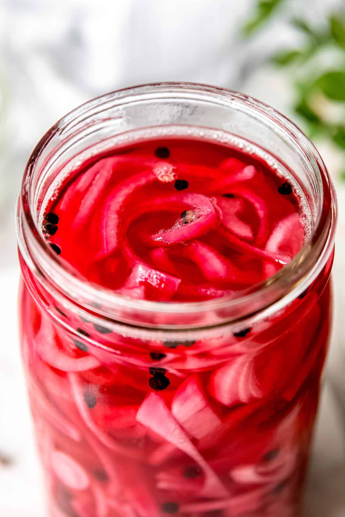 45 degree angle closeup on a jar of quick pickled red onions; you can see the vibrant color of the onions and brining liquid with whole peppercorns floating in the brine.
