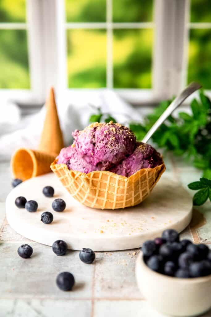 waffle cone bowl of blueberry cheesecake ice cream on a white marble round on a grey slate tile table with a pinch bowl of fresh blueberries in the foreground with blueberries scattered around a couple of ice cream cones in the background.