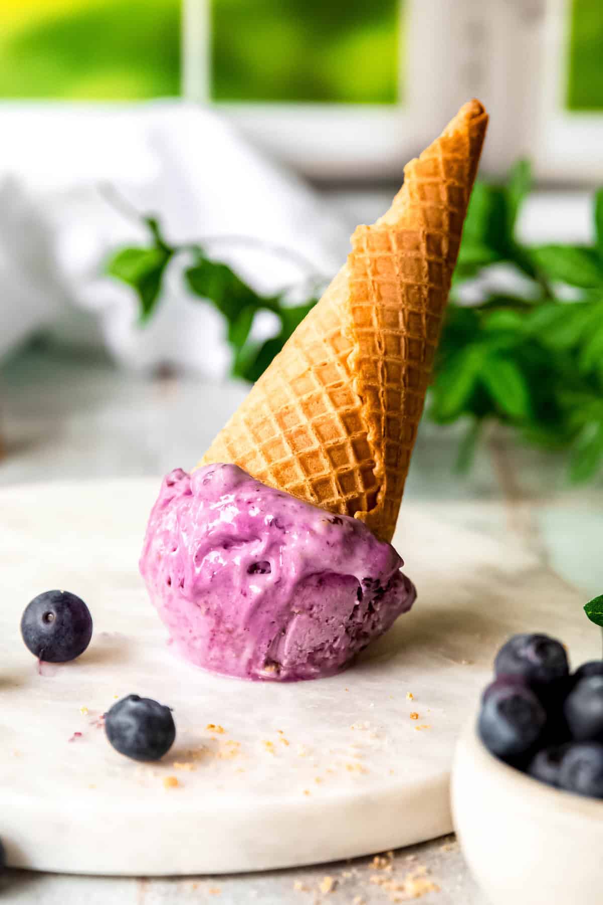 blueberry ice cream cone flipped on its head on a white marble round with fresh blueberries and graham cracker crumbles scattered around.