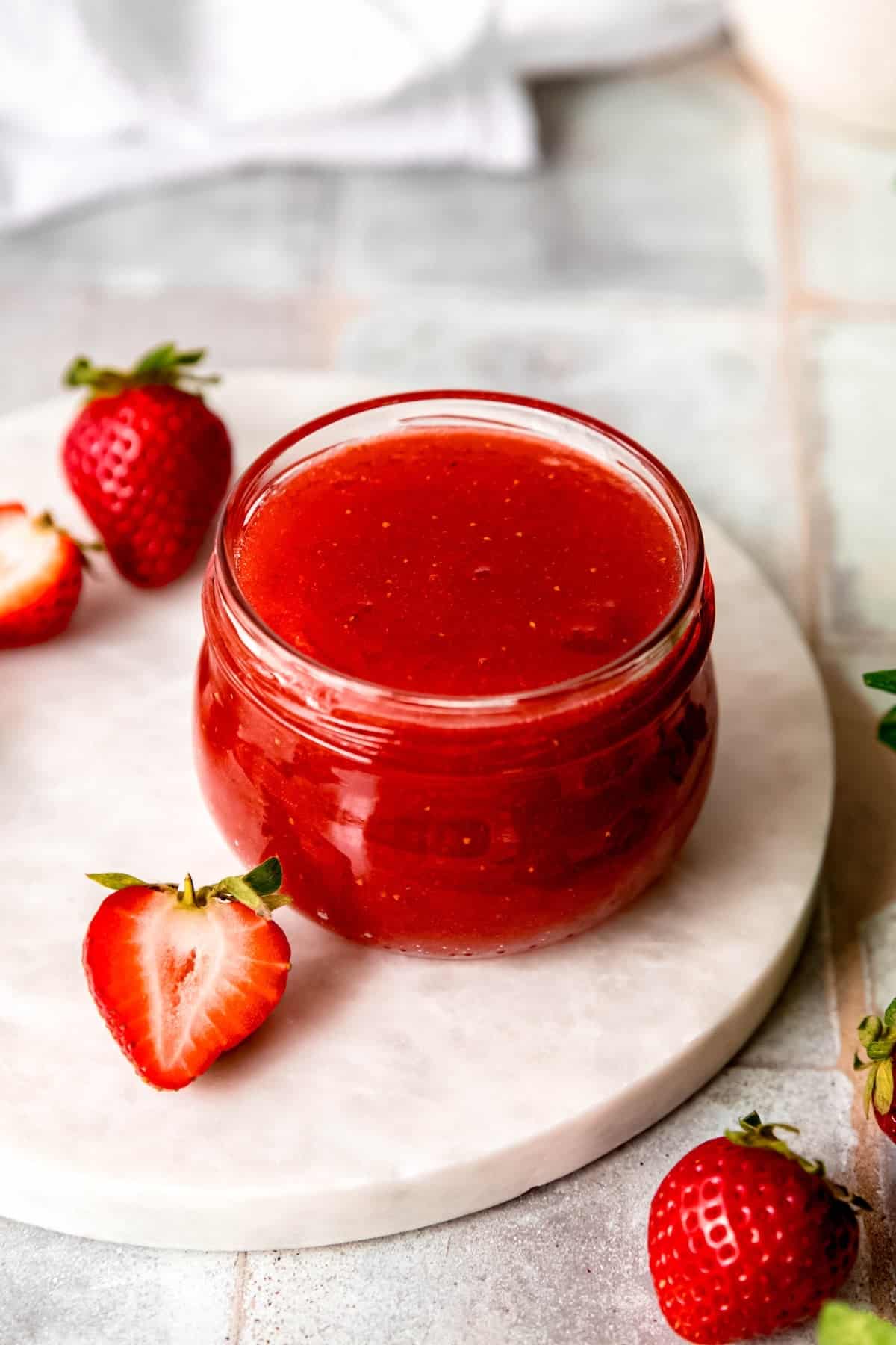45 degree angle shot of a jar of canned strawberry jam on a white table with fresh strawberries.