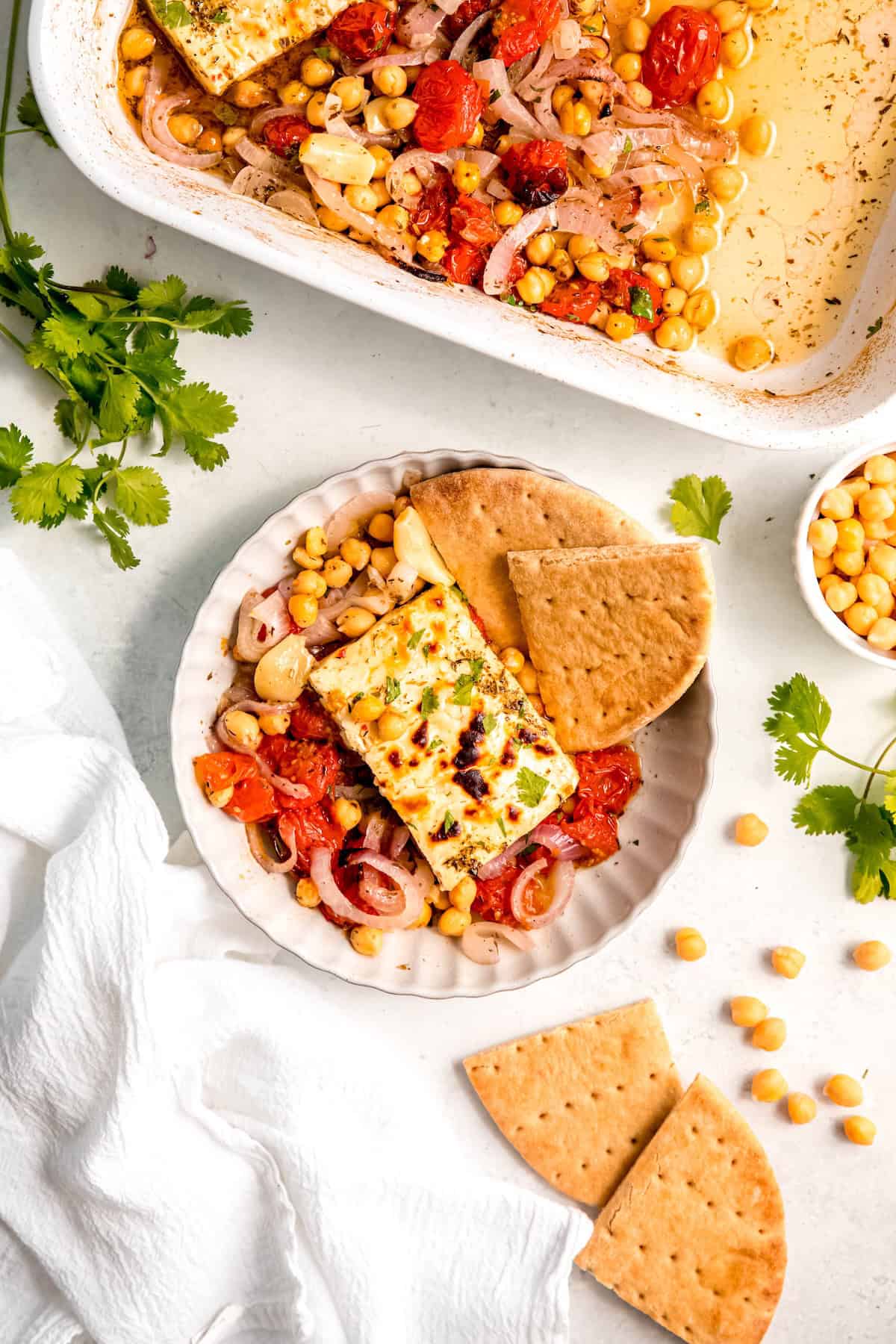 flat lay shot of a serving of Mediterranean baked feta with tomatoes and chickpeas in a white pasta bowl on a table with fresh cilantro, a pinch bowl of chickpeas, and several wedges of pita bread.