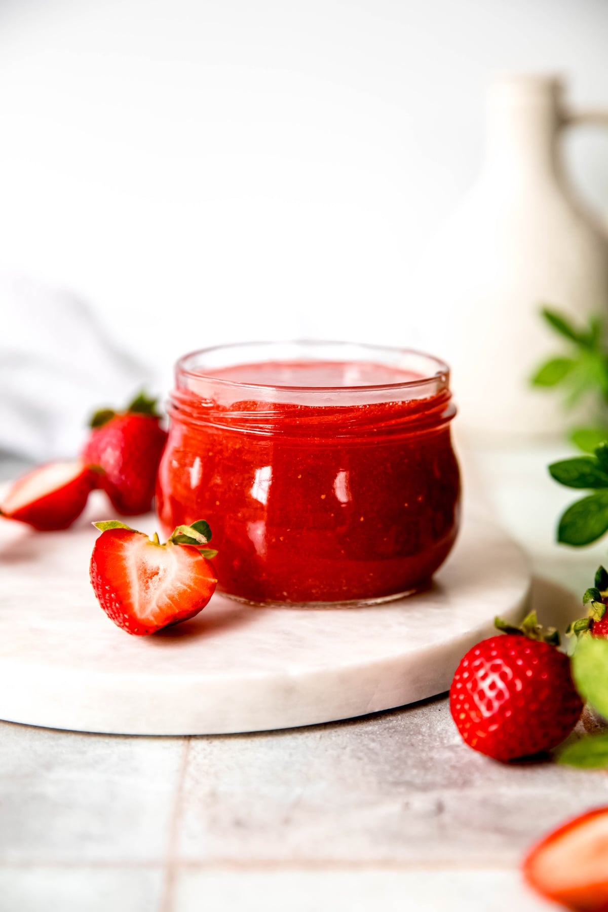 half pint mason jar filled with homemade strawberry jam with pectin on. a white table with whole and halved strawberries and a green houseplant.