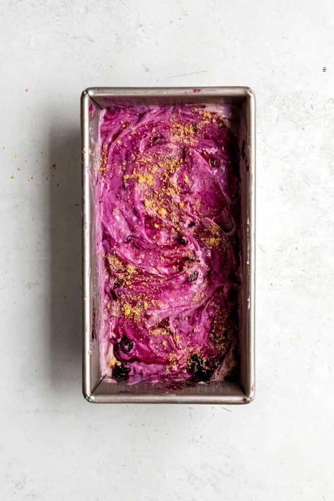 blueberry cheesecake ice cream in the loaf tin after adding multiple layers of ice cream, blueberry sauce, and graham cracker crumbles.