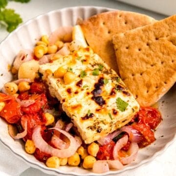 45 degree closeup of a piece of Mediterranean baked feta on a bet of roasted tomatoes and chickpeas with two wedges of pita bread.