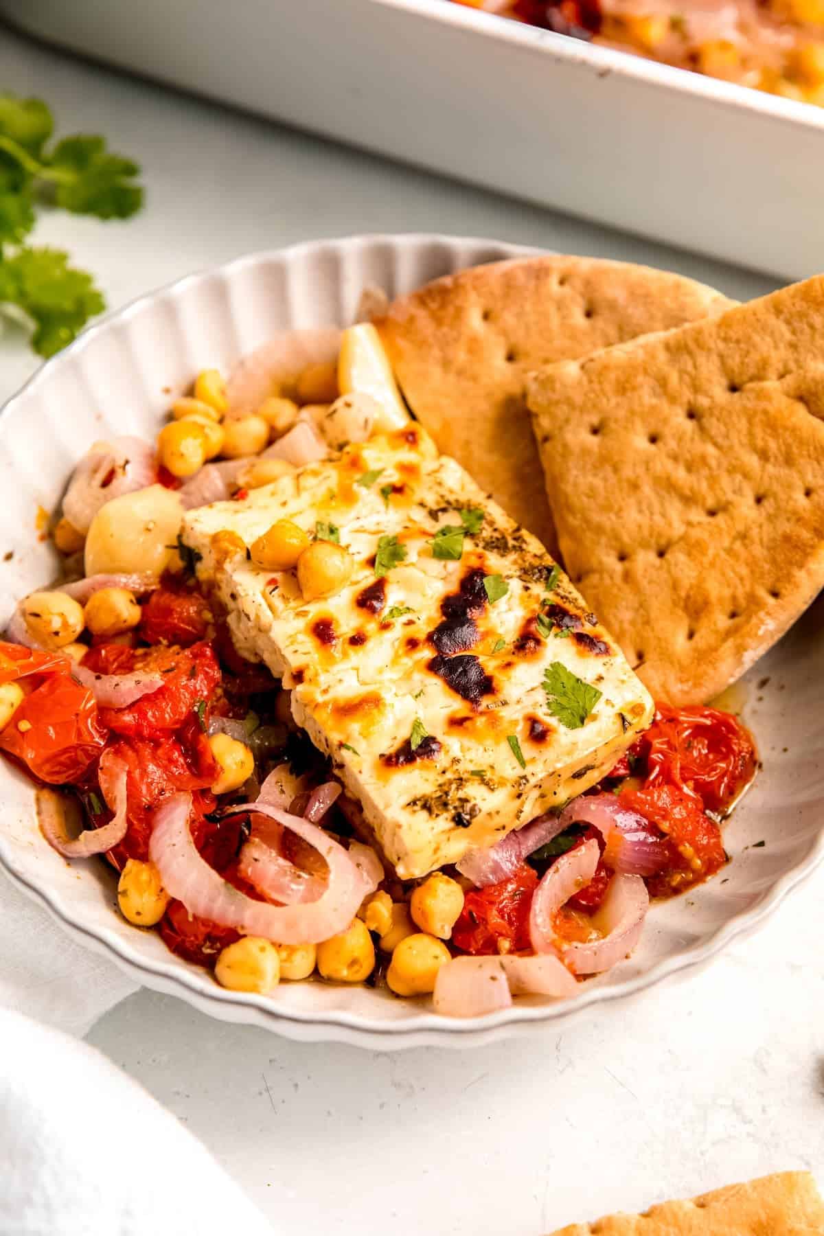 45 degree closeup of a piece of Mediterranean baked feta on a bet of roasted tomatoes and chickpeas with two wedges of pita bread.