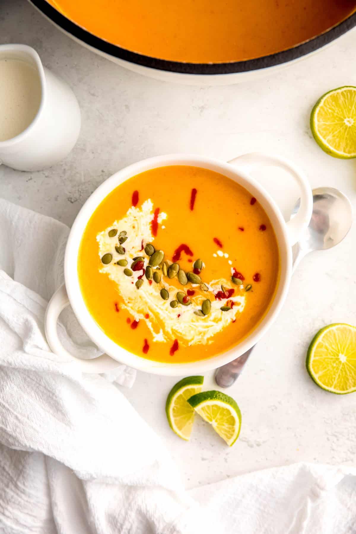 flat lay shot of a white soup bowl filled with vibrant spicy apple and butternut squash soup with all the fixins—a drizzle of coconut milk, sriracha, and pepitas, with lime wedges on the side for serving.