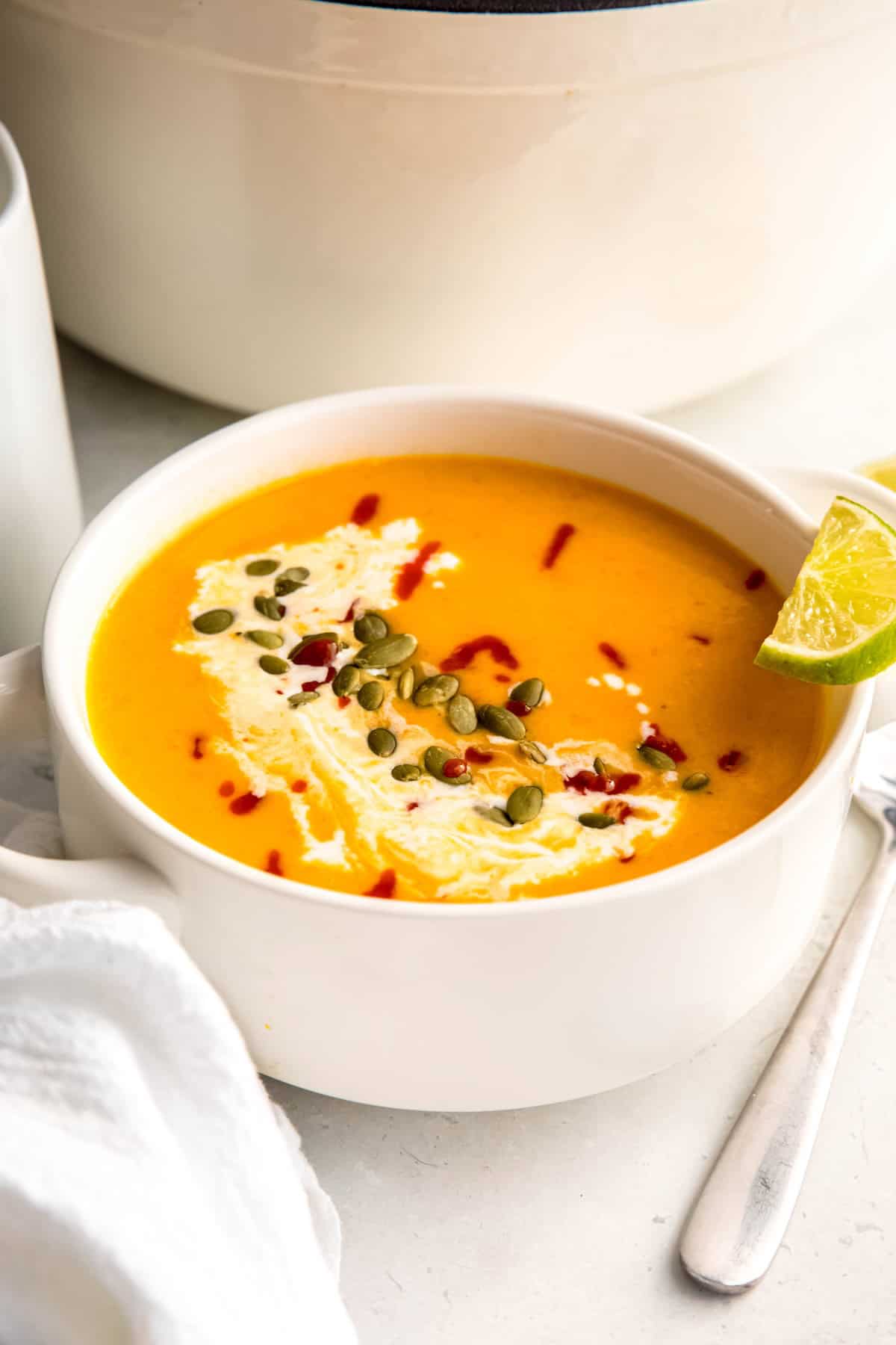 45 degree angle closeup of a bowl of spicy apple butternut squash soup topped with pepitas, coconut milk, and sriracha with a lime wedge perched on the side.