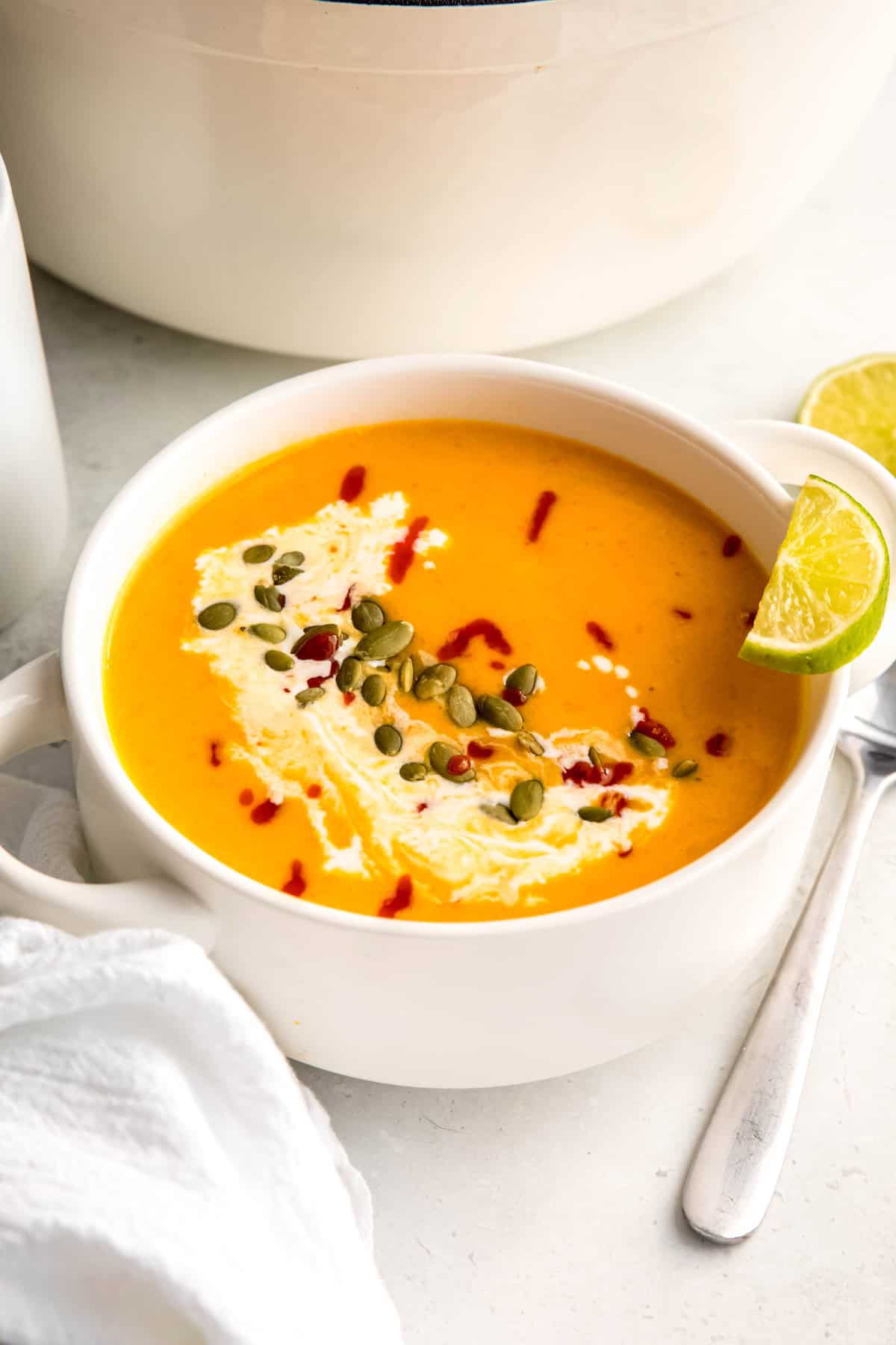 45 degree angle of a white soup bowl filled with spicy butternut squash and apple soup topped with pepitas, sriracha, coconut milk, and a lime wedge in front of a white dutch oven on a white table with a white linen napkin and silver soup spoon.