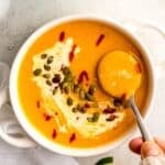 silver spoon taking a bite of spicy butternut squash curry soup topped with pumpkin seeds, sriracha, and coconut cream.