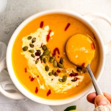 silver spoon taking a bite of spicy butternut squash curry soup topped with pumpkin seeds, sriracha, and coconut cream.