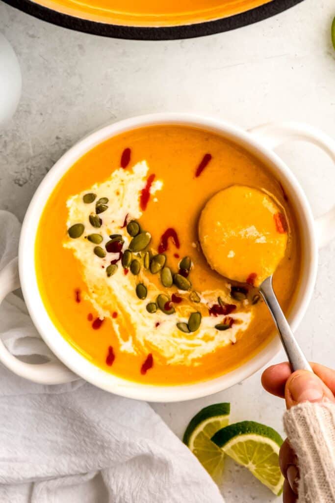 silver spoon taking a bite of spicy butternut squash curry soup topped with pumpkin seeds, sriracha, and coconut cream.
