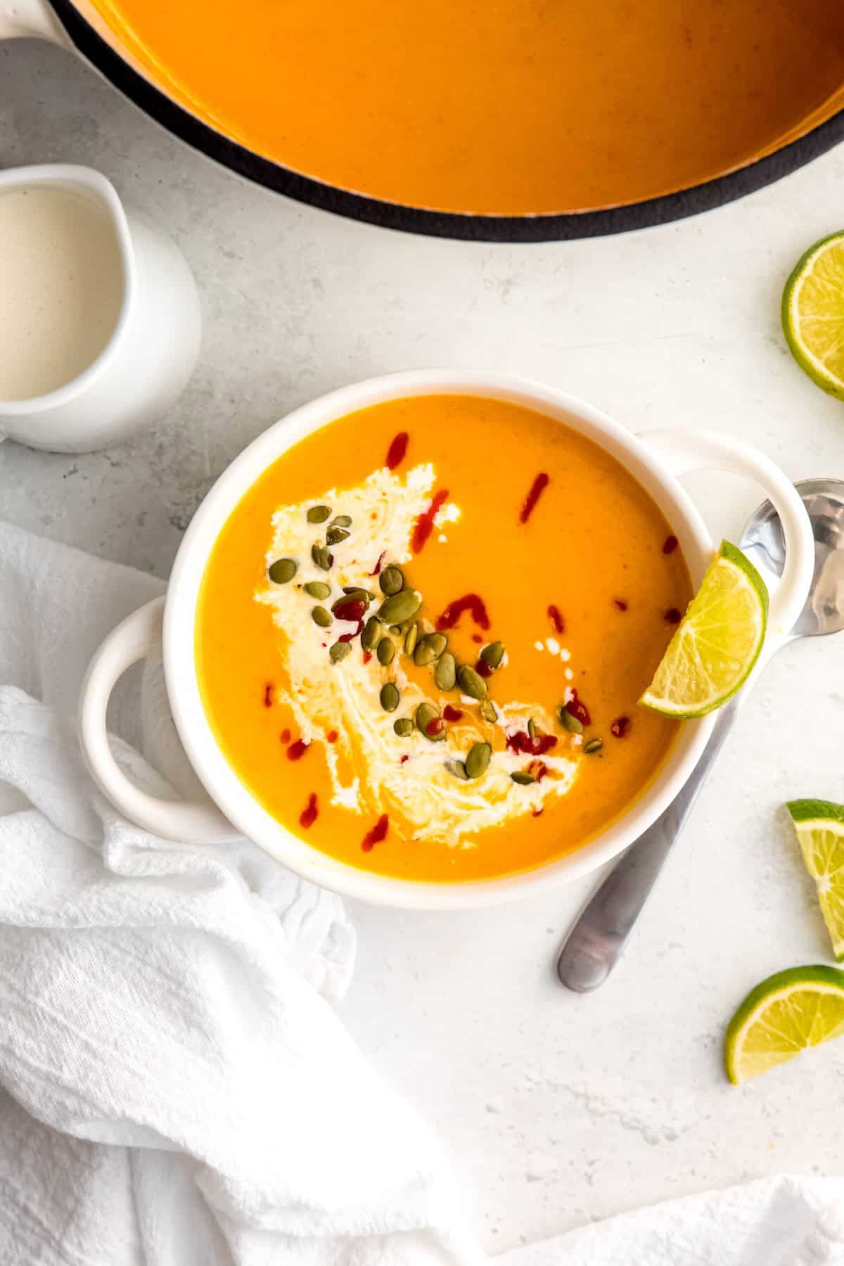 overhead shot of a white soup bowl with handles filled with a portion of spicy butternut squash and apple soup topped with pepitas, hot sauce, and coconut milk with a sliced lime on the side.