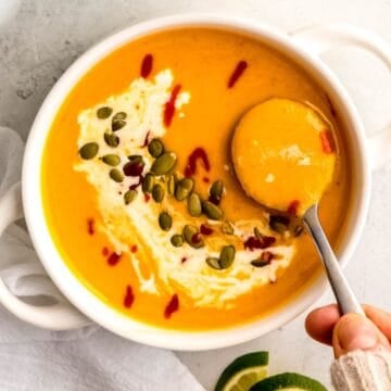 square hero image of a hand in a white sweater up to the knuckles taking a spoonful of spicy butternut squash soup.