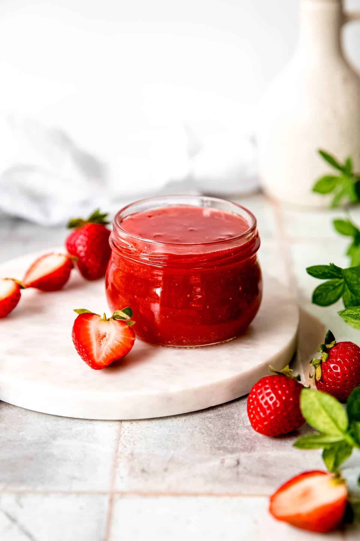 half pint mason jar of sure jell strawberry jam on a white table with fresh strawberries and a green houseplant.