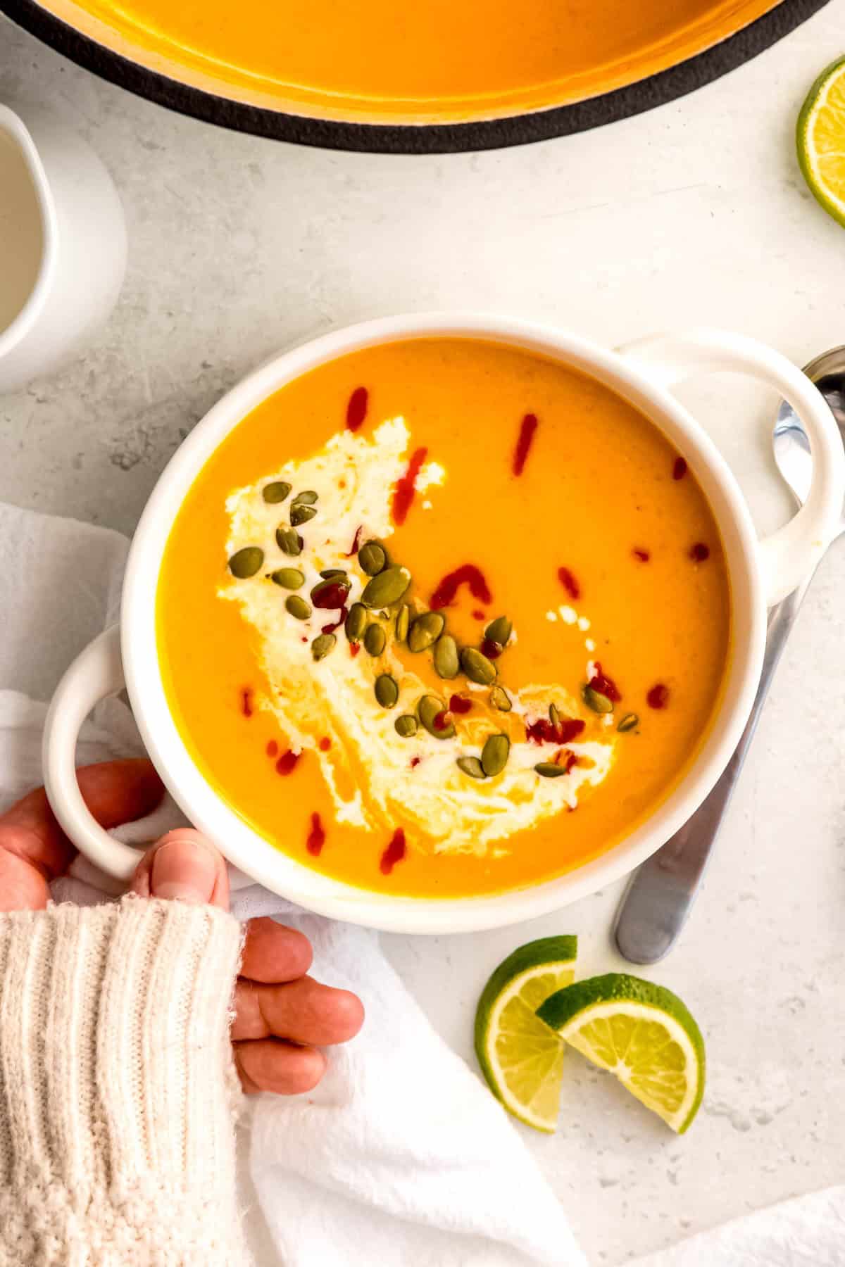 hand clad in a cozy white sweater up to the knuckles setting down a bowl of thai inspired spicy butternut squash soup down on a white table with a silver soup spoon and lime wedges.