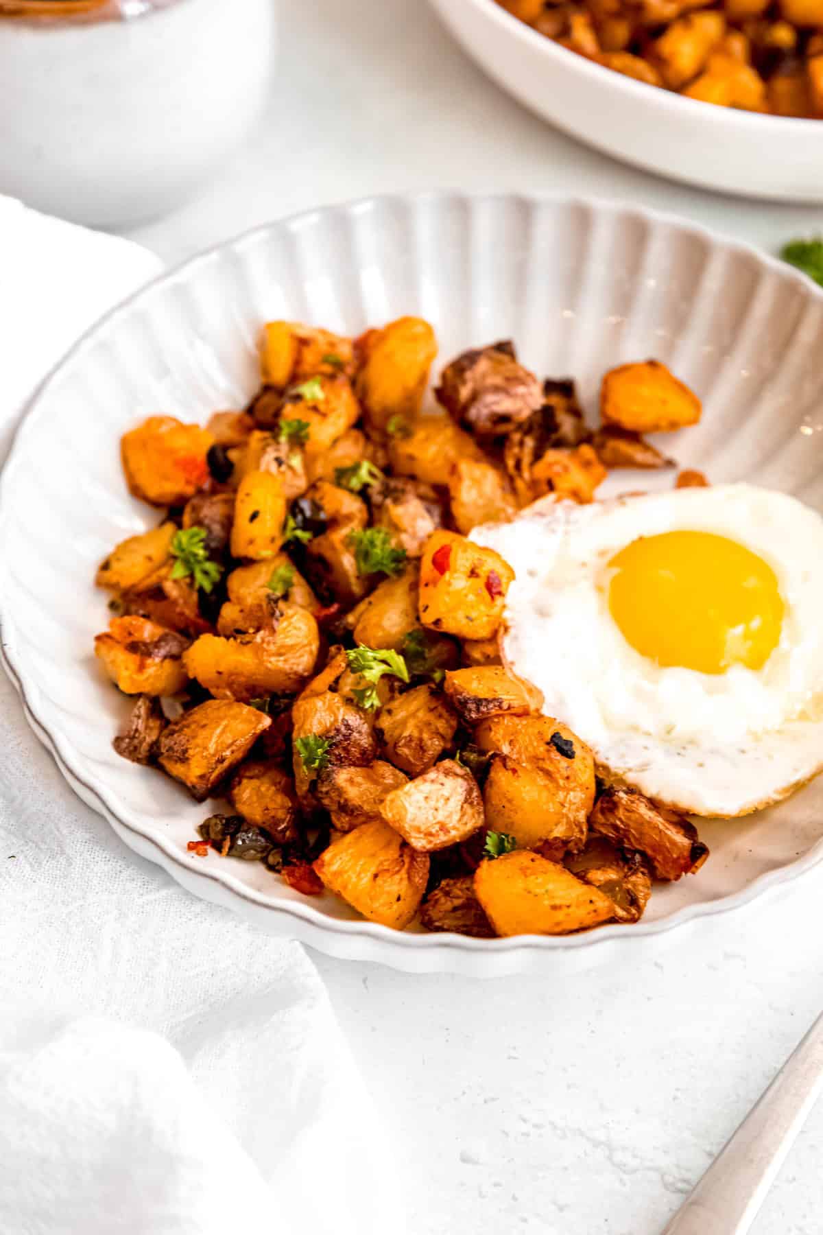 serving of baked home fries, onions and peppers in a white scalloped bowl with a fried egg and a silver fork.