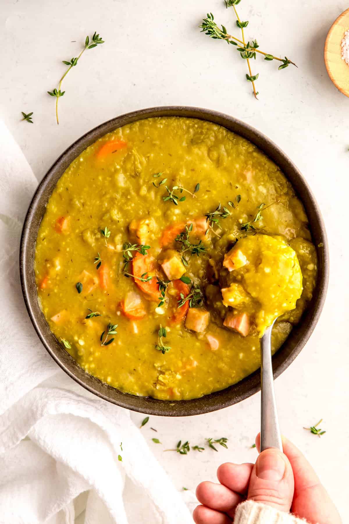 hand clad in a white sweater up to the knuckles holding a silver spoonful of chunky ham and split pea soup above the bowl.